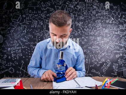 Hipster insegnante presso la scrivania con microscopio, big blackboard Foto Stock