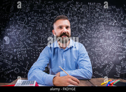 Felice maestro seduto alla scrivania, forniture scolastiche, big blackboard Foto Stock