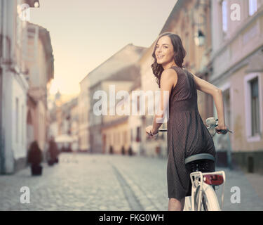 Bella giovane donna con la bicicletta ferma a girare la testa e ci danno un sorriso toothy alla fotocamera in una città europea al tramonto Foto Stock