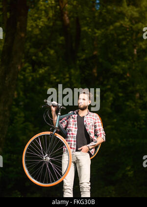 Il blue-eyed bel giovane vestito casual bicicletta sulle spalle e schiena verde della foresta Foto Stock