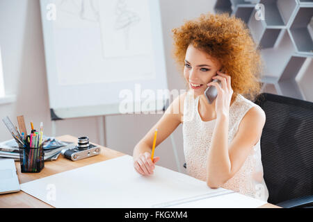 Allegro bella donna giovane fotografo seduto sul posto di lavoro e parlando al telefono cellulare Foto Stock