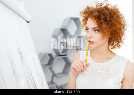 Malinconici affascinante redhead giovane donna fare schizzi e pensare in ufficio Foto Stock