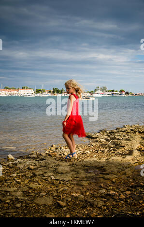 Bella ragazza bionda in abito rosso camminando lungo la riva del mare Foto Stock