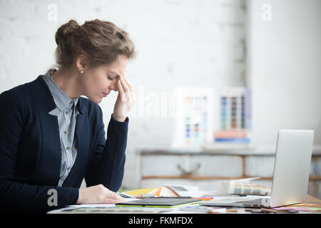 Ritratto di giovane donna ha sottolineato seduti a casa office desk nella parte anteriore del computer portatile, toccando la testa con stanco espressione facciale Foto Stock