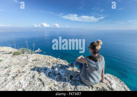 Vista posteriore del giovane donna seduta sulla scogliera che guarda a vista oceano, Cagliari, Sardegna, Italia Foto Stock