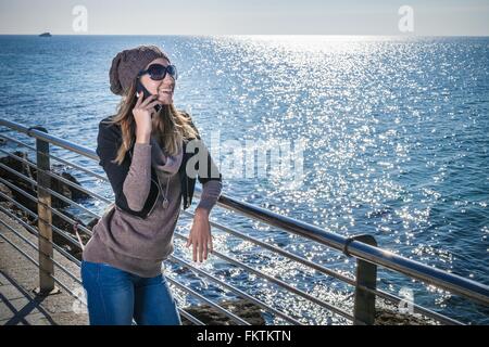 Giovane donna appoggiata contro le ringhiere dall' oceano utilizza lo smartphone che guarda lontano sorridente Foto Stock