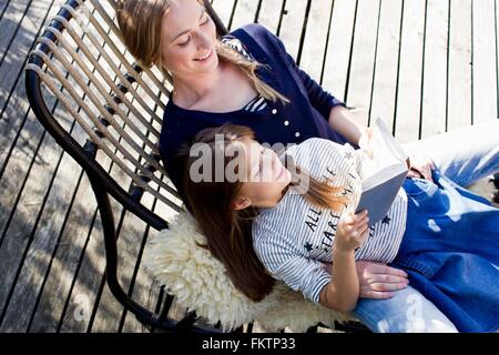 Madre e figlia sul rilassante lounge chair, lettura della ragazza prenota Foto Stock