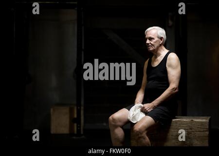 Senior uomo seduto appoggiato in palestra scuro Foto Stock