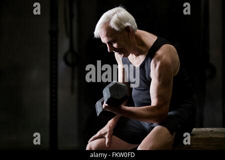 Senior uomo seduto facendo bicipite ricci con il manubrio in palestra scuro Foto Stock