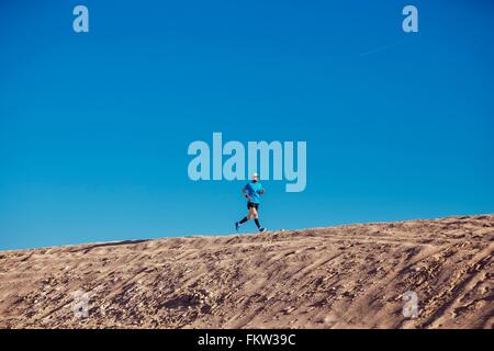 Basso angolo vista del cielo blu e metà uomo adulto in esecuzione sulla duna di sabbia, Dubai, Emirati Arabi Uniti Foto Stock