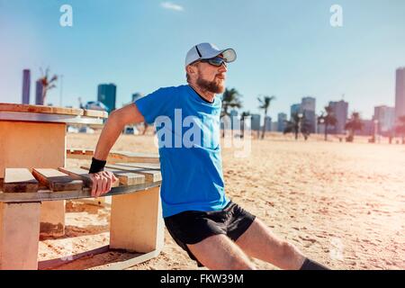 Metà uomo adulto sulla spiaggia facendo retromarcia push up su banco, Dubai, Emirati Arabi Uniti Foto Stock