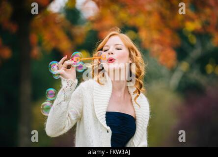 Giovane donna bella con il rosso dei capelli ondulati soffiare bolle nella foresta di autunno Foto Stock