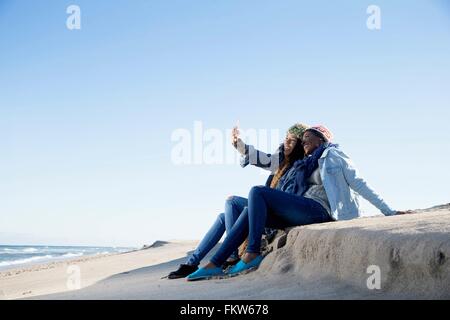 Due amici seduti sulla spiaggia, tenendo autoritratto, utilizza lo smartphone Foto Stock