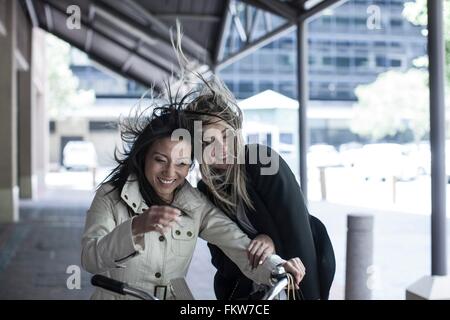 Due amici delle donne con capelli oltremare ridere in bicicletta in città Foto Stock