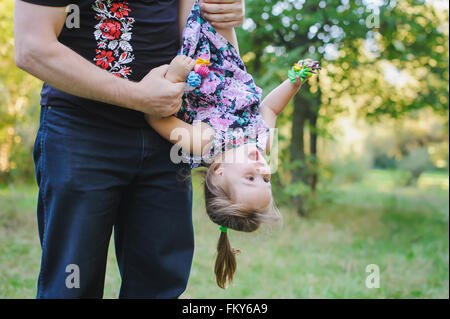 Papà gioca con il suo piccolo felice figlia, tenendo il suo capovolto in un verde prato Foto Stock