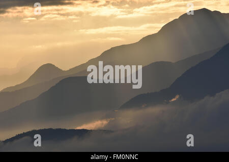 Misty creste montane all'alba con soffici nuvole immerso nelle valli, illuminata dai raggi del sole. Foto Stock