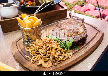 32oz tomahawk bistecca, patatine e cipolle di tabacco, presentato su un tagliere di legno e cesto in filo. Foto Stock