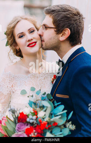 Felice lo sposo bacia dolcemente la sua splendida bionda sposa su sfondo bianco close up Foto Stock