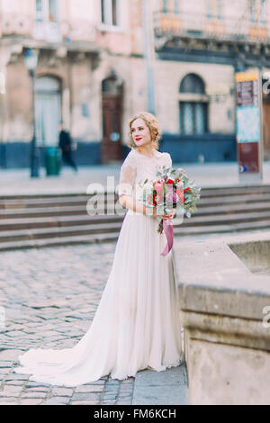 Affascinante bionda sposa allegramente sorridente con bouquet in mani e antica architettura di Lviv sullo sfondo Foto Stock