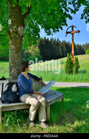 Donna seduta su un banco di lavoro sotto un albero, croce sul ciglio della strada nei pressi di Ölmühle in Rotenbachtal Valley, vicino a Ellwangen, Baden-Württemberg Foto Stock