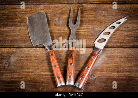 Acciaio inox Coltello per formaggio, forcella e Cleaver con manico di legno sono distesi sulla scrivania in legno. Foto Stock