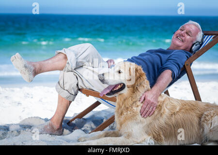 Uomo maturo in appoggio su una sedia a sdraio ascoltando la musica come accarezzare il suo cane Foto Stock