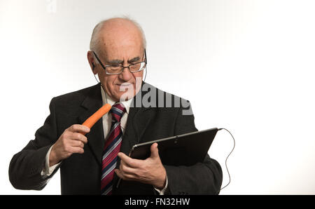 Vecchio imprenditore senior lavora con nero computer tablet, indossando il business suit, mangiando la carota Foto Stock