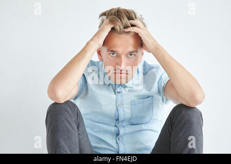 Uomo brutale in una camicia con maniche corte seduta sul pavimento, sotto lo sguardo della fronte, tenendo la sua testa Foto Stock