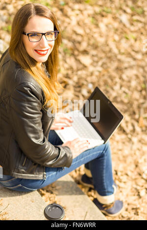 Giovane donna felice con capelli lunghi utilizzando laptop su scale nel parco. Vista aerea dell'immagine. Calda tonalità di colore immagine Foto Stock