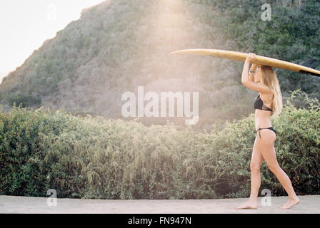 Una donna in bikini nero a piedi giù per un sentiero che porta una tavola da surf sulla sua testa. Foto Stock