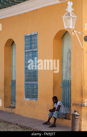 La vita quotidiana a Cuba - afro-caraibica uomo seduto sulla soglia dell edificio di colore luminoso guardando in giù a Trinidad, Cuba Foto Stock
