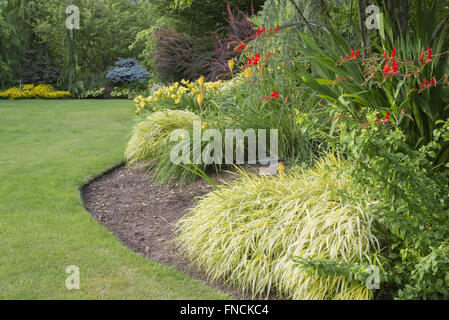 Un lussureggiante verde prato dolcemente curve intorno a una grande varietà di alberi e fiori perenni, come daylilies e rosso brillante crocosmia con Foto Stock