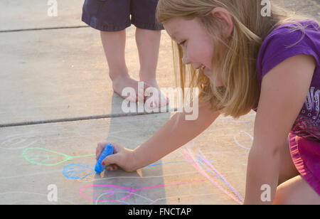 Una giovane ragazza di disegno con gesso su una luce colorata vialetto mentre suo fratello più giovane orologi. Foto Stock