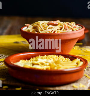 Primo piano di una piastra di terracotta con un po' di pasta cruda e un vaso di terracotta con spaghetti alla carbonara, su un tavolo Foto Stock