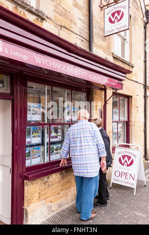 Un elderley giovane guardare in un agente immobiliare nella finestra pretty Cotswold città di Burford , Oxfordshire , Inghilterra , Inghilterra , Regno Unito Foto Stock