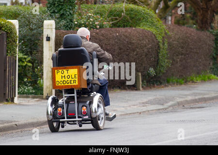 Un utente in sedia a rotelle a Tarleton, Lancashire, Regno Unito. 16 marzo 2016. Esponente senior e pensionato che mostra un cartello che evidenzia lo stato delle strade del Lancashire per i veicoli scooter a mobilità motorizzata. Questo tratto è così male gli utenti di sedie a rotelle e scooter di mobilità devono viaggiare su strada. Gli scooter da 6-8 miglia/h Mobility a causa della loro maggiore velocità devono essere guidati su strada, poiché la legge vieta lo spostamento dello scooter a una velocità di 4 miglia/h sul marciapiede, mentre i carrelli più lenti sono limitati a, e costretti a sopportare un viaggio sconnesso sul marciapiede. Foto Stock