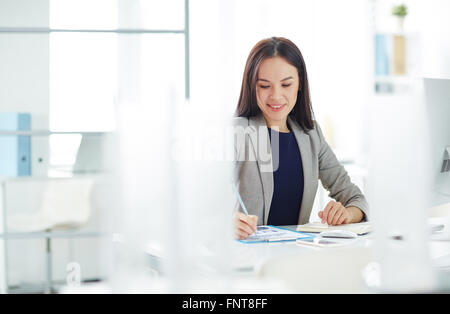 Sorridente giovane imprenditrice seduta sul suo posto di lavoro e il lavoro con i documenti Foto Stock