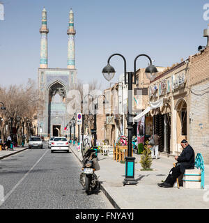 Cityscape, Yazd, Iran. Un uomo anziano si siede su un banco di lavoro come gli altri andare circa la loro attività. Moschea del Venerdì visibile alla fine della strada. Foto Stock