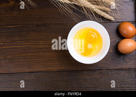 Uova fresche con due rotte in una ciotola visualizzando il giallo tuorli pronti per la cottura su una tavola di legno con spighe di grano e copia dello spazio. Foto Stock