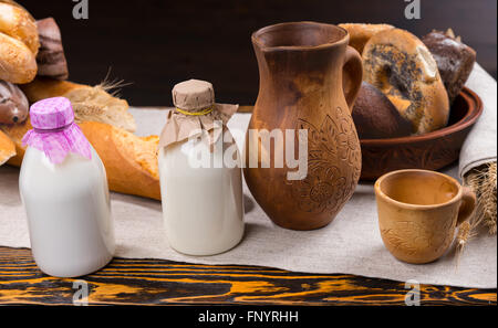 Due piccole bottiglie di latte con carta copertine accanto al lanciatore di legno, coppa e varie forme di pane e panini di pane sul tavolo Foto Stock