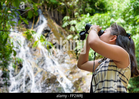 Bellissima fanciulla escursionismo è utilizzando un binocolo per guardare gli uccelli nella foresta tropicale vicino alle cascate in Thailandia Foto Stock