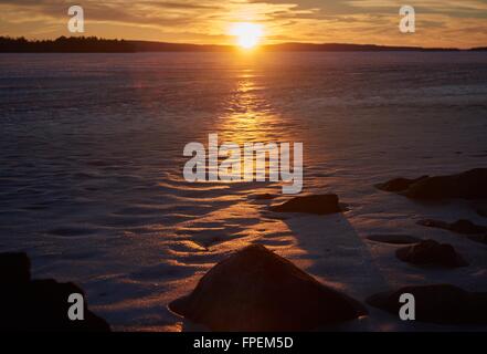 Tramonto a un lago ghiacciato in inverno sera in Finlandia. I colori caldi del sole al tramonto sul ghiaccio freddo. Foto Stock