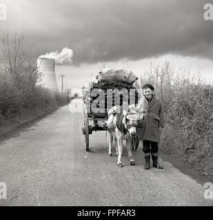 Storia degli anni '50, l'immagine mostra un giovane irlandese su una corsia di campagna con un vecchio carrello con ruote in legno pieno di erba sintetica o torba tirata da un piccolo asino, l'Irlanda. Foto Stock