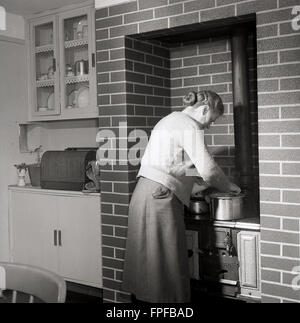 1950s storico, una signora al suo fornello o gamma, costruito in un camino di mattone di camino circondare, controllando su una pentola di metallo, Irlanda. Quando le stufe rimpiazzarono fuochi aperti per riscaldare una proprietà, modelli furono sviluppati che potevano anche essere usati per cucinare e questi furono riferiti come intervalli, come molti avevano più di un forno e due punti di cottura sulla parte superiore, dando una 'gamma' di posti su cui cucinare. Foto Stock