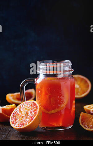 Tagliate a fette il sangue siciliano arance e vetro mason jar di rossi freschi succhi di arancia sopra il vecchio tavolo in legno. Scuro in stile rustico. Foto Stock