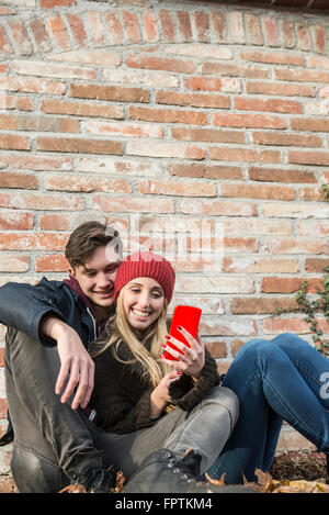 Coppia giovane sorridente e utilizza lo smartphone contro un muro di mattoni, Monaco di Baviera, Germania Foto Stock