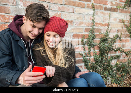 Coppia giovane sorridente e utilizza lo smartphone contro un muro di mattoni, Monaco di Baviera, Germania Foto Stock