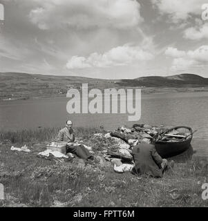 Storico degli anni '50, foto del fotografo inglese J Allan Cash fare un picnic accanto ad un lago, Irlanda, con un uomo locale per compagnia. Una piccola barca a remi in legno è ormeggiata al bordo dell'acqua. Foto Stock