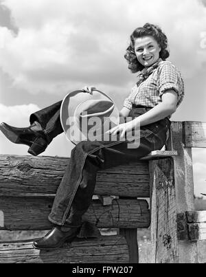 1940s sorridente BRUNETTE donna che indossa jeans WESTERN USURA tenendo il cappello da cowboy seduto sulla staccionata in legno guardando la fotocamera Foto Stock