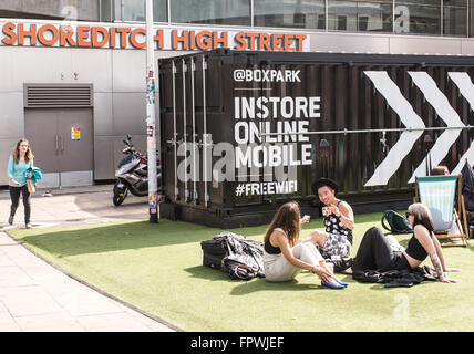 Gruppo di giovani hipsters seduti su un finto tappeto d'erba in boxpark, il primo al mondo pop-up mall basato in Shoreditch. Foto Stock